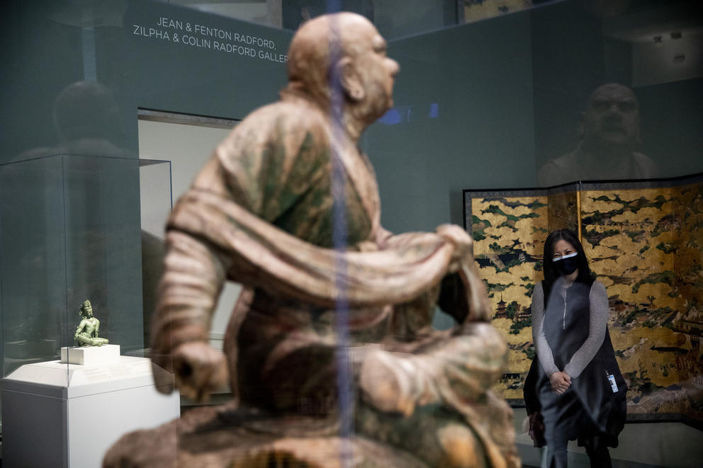 Image of a brown sculpture of a bald man wrapped in cloth, sculpture is sitting in a glass display case. in the distance, a woman with a mask in front of a painted panel looks on.