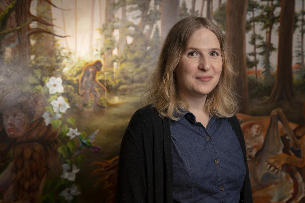 Person with blonde hair in dark clothes poses in front of a mural that shows lush woods and a sasquatch-like animal walking