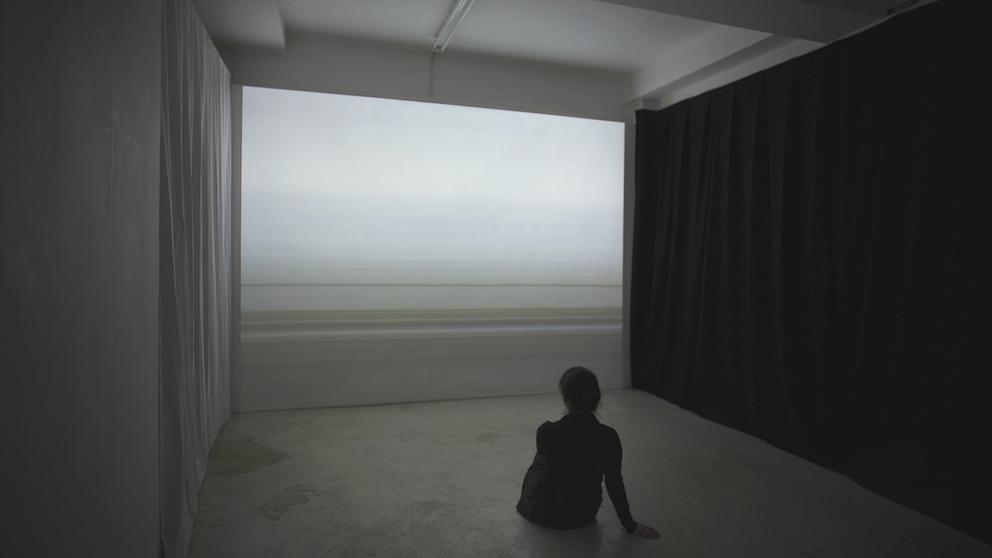 A woman sitting on concrete floor looking out a wall-sized window into dull ocean/sky horizon