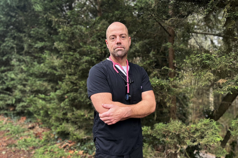 Nurse Steven Higgs poses with his arms crossed in dark blue scrubs in front of evergreen trees. 