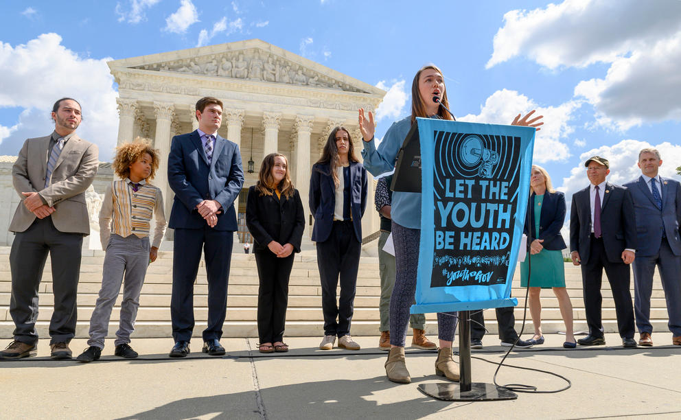 A group of youth plaintiffs stand in front of the Supreme Courthouse in suits, with one young woman giving a speech behind a teal poster.