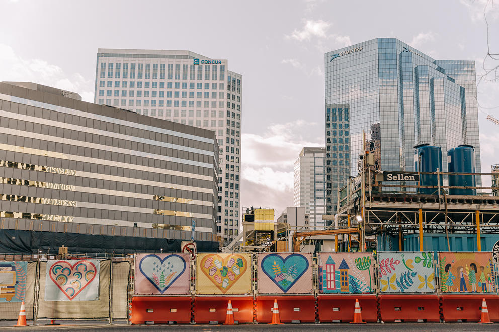 Tall shiny buildings rise above a construction fence with colorful heart-shaped artwork