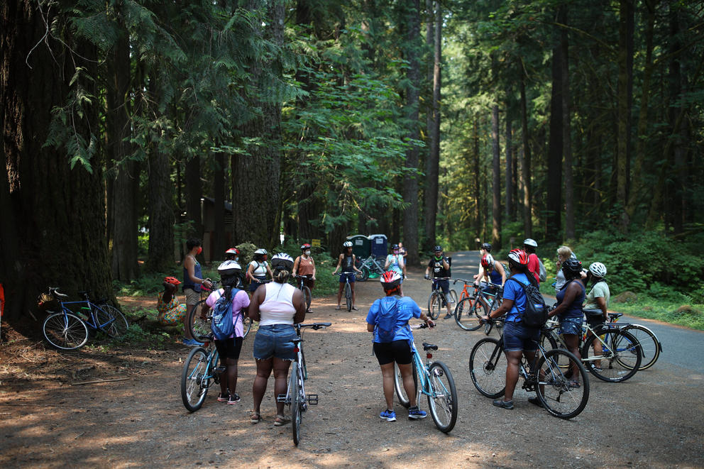 Over a dozen cyclists stand in a circle with their bikes in a forest