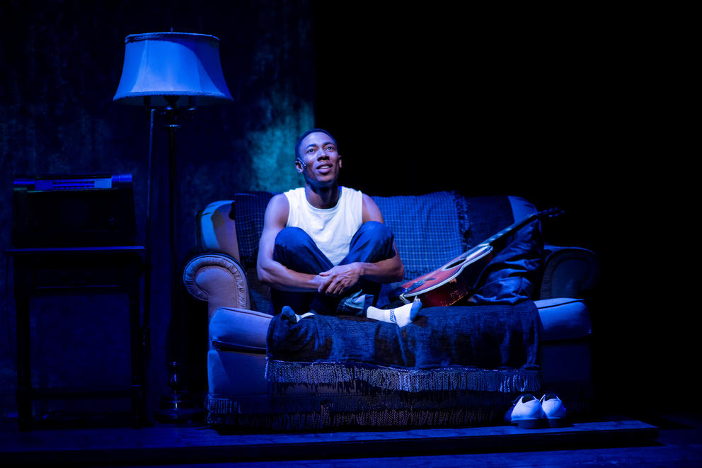 photo of an actor on stage - a boy in a white t-shirt sitting on a couch with a guitar