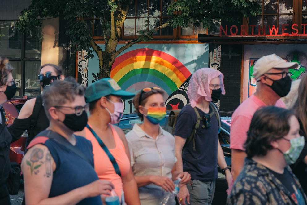 a group of people wearing facial masks walks together in front of a large rainbow mural