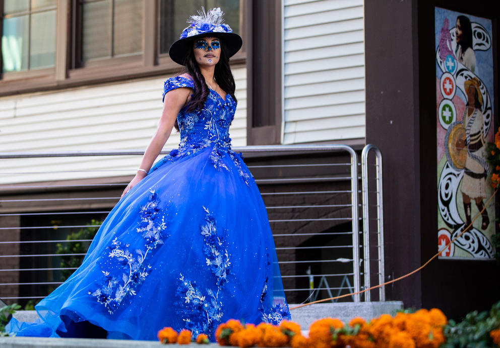 A woman in an elaborate blue dress and hat wears Day of the Dead face paint