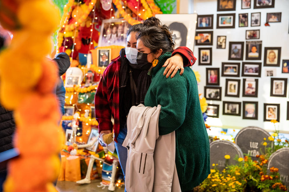 two women wearing masks hug, behind them a wall is lined with photographs of people and orange decorations