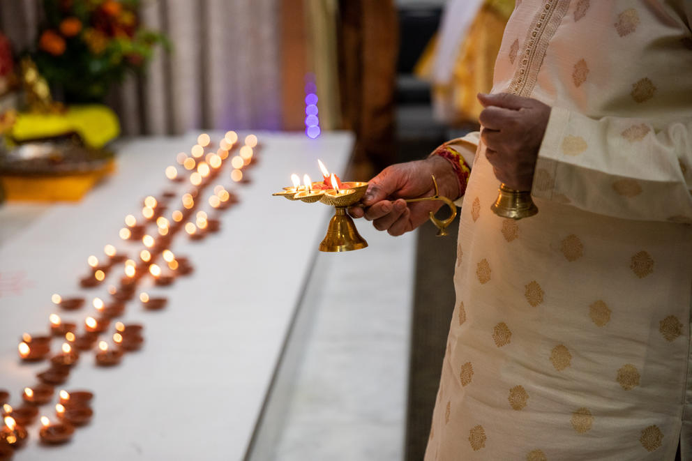 A hand holds a gold holder with candles over a stage with row of clay bowls lit with flames