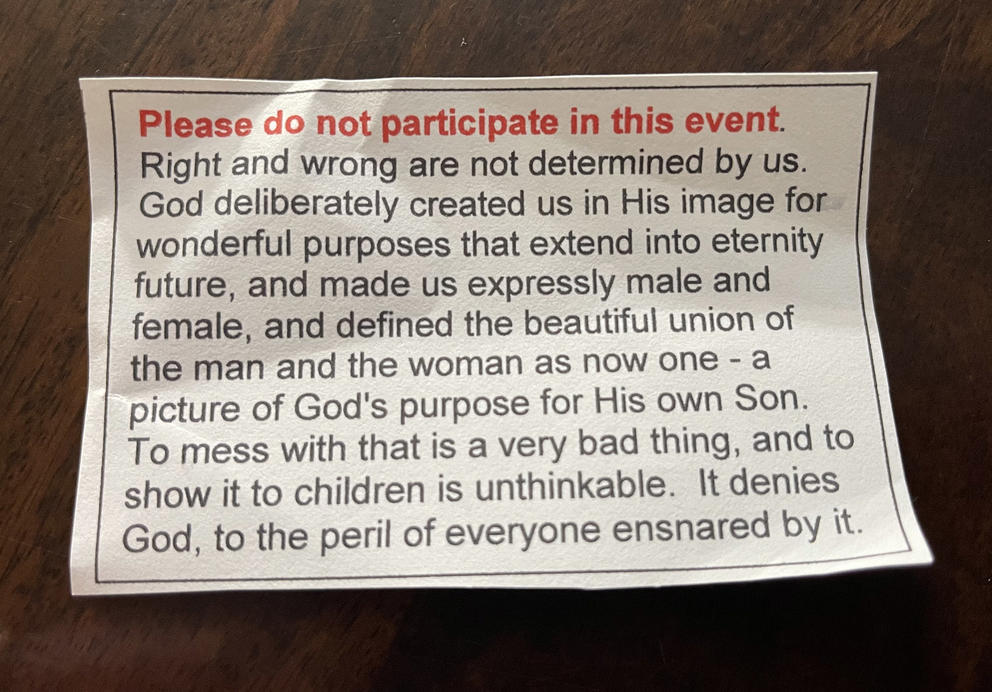 Notecard reads: "Please do not participate in this event. Right and wrong are not determined by us. God deliberately created us in His image for wonderful purposes that extend into eternity future, and made us expressly male and female, and defined the beautiful union of the man and the woman as now one – a picture of God's purpose for His own Son. To mess with that is a very bad thing, and to show it to children is unthinkable. It denies God, to the peril of everyone ensnared by it."