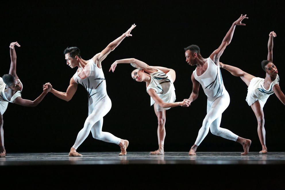 Dancers in white clothing are dancing on a dark stage