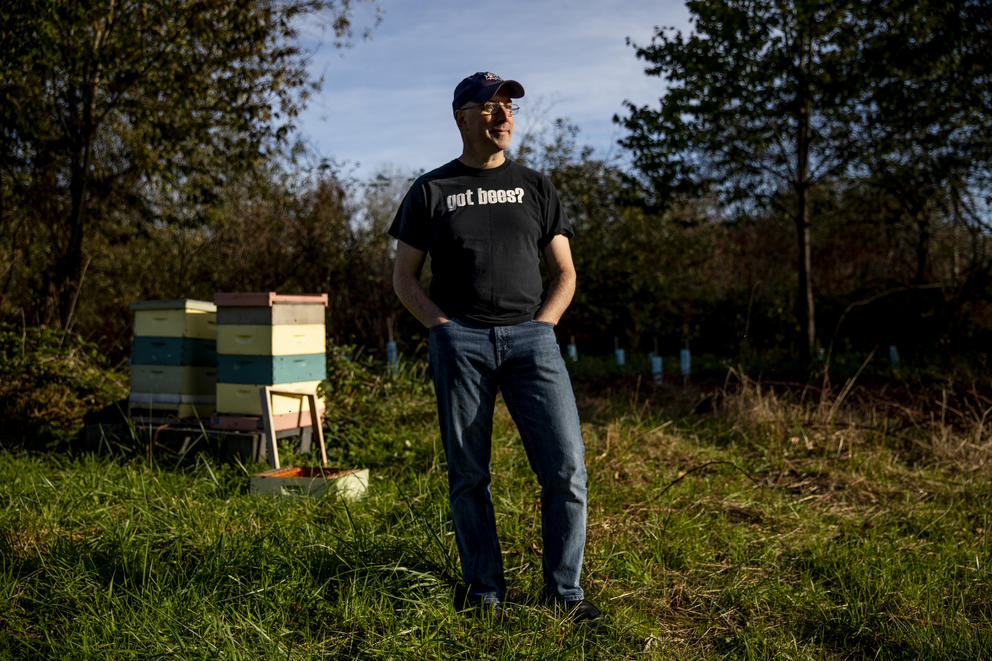 A man stands in an orchard with a beehive behind him