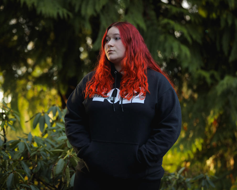 A woman with bright red hair stands outside in front of trees lit up by evening sun