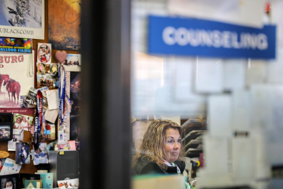 A woman is seen through a window of a school Counseling office, photos and keepsakes are posted to the walls
