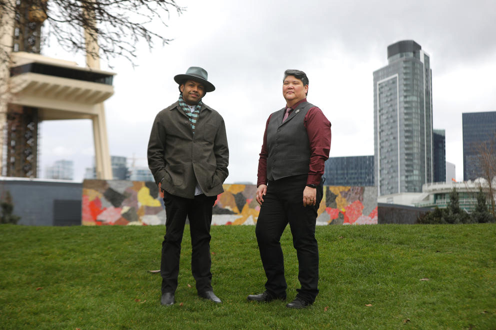 Northwest Folklife co-directors Reese Tanimura and Benjamin Hunter stand on a grassy hill with the partial downtown Seattle skyline and Space Needle behind them 