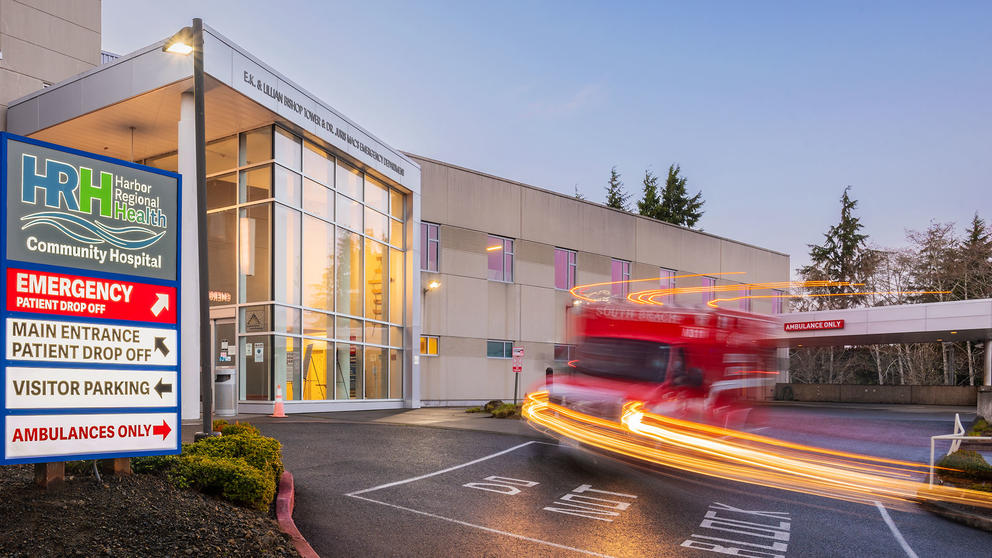 A long-exposure photo depicts an ambulance streaking past the front windows of the Harbor Regional Health emergency department.
