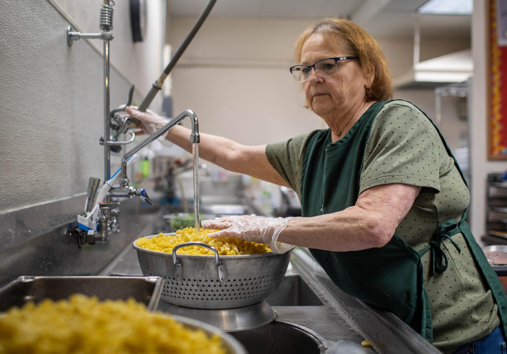 Cathy Rupe leans over a sink, holding a large bowl of macaroni and cheese in a large elementary school kitchen.