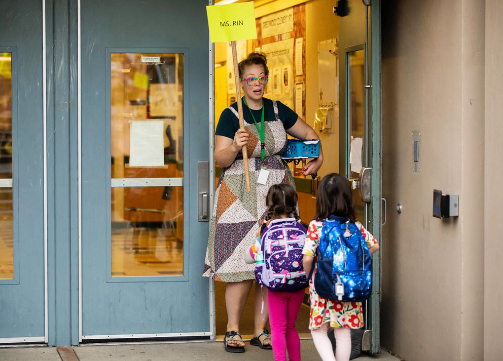 A teacher holding a sign that says "Ms. Rin" stands at a door of a school, as two students file in.