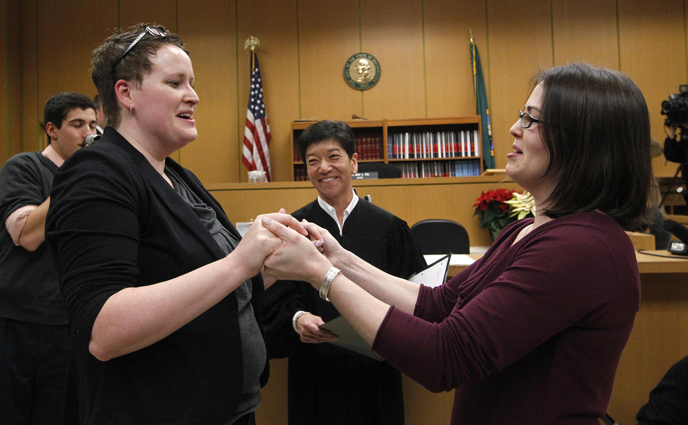 Two women hold hands as they get married, the judge smiles between them