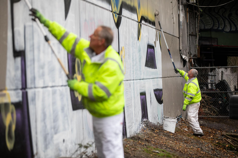 Two men in white paints and hi-vis jackets use rollers to paint over graffiti