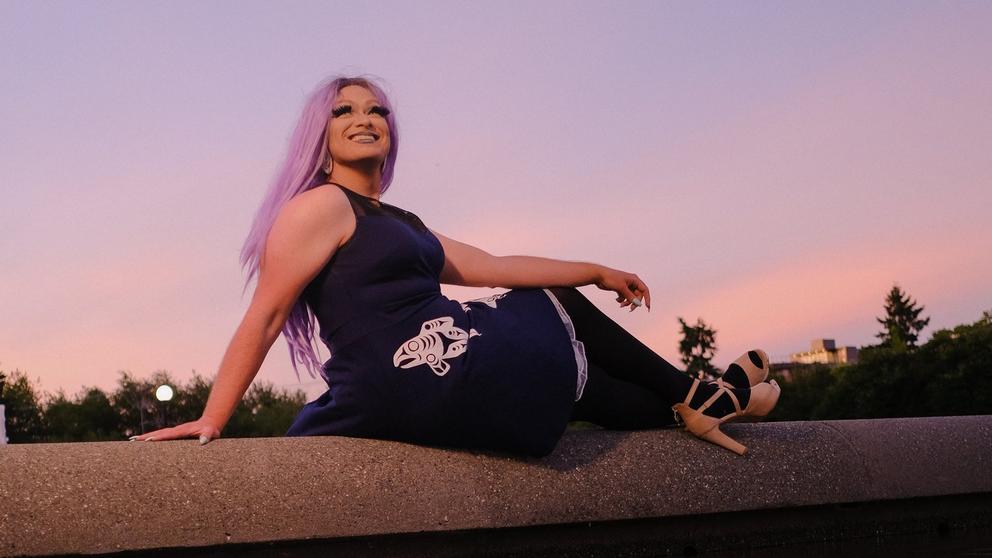 a woman with long pink hair sits on a wall at sunset