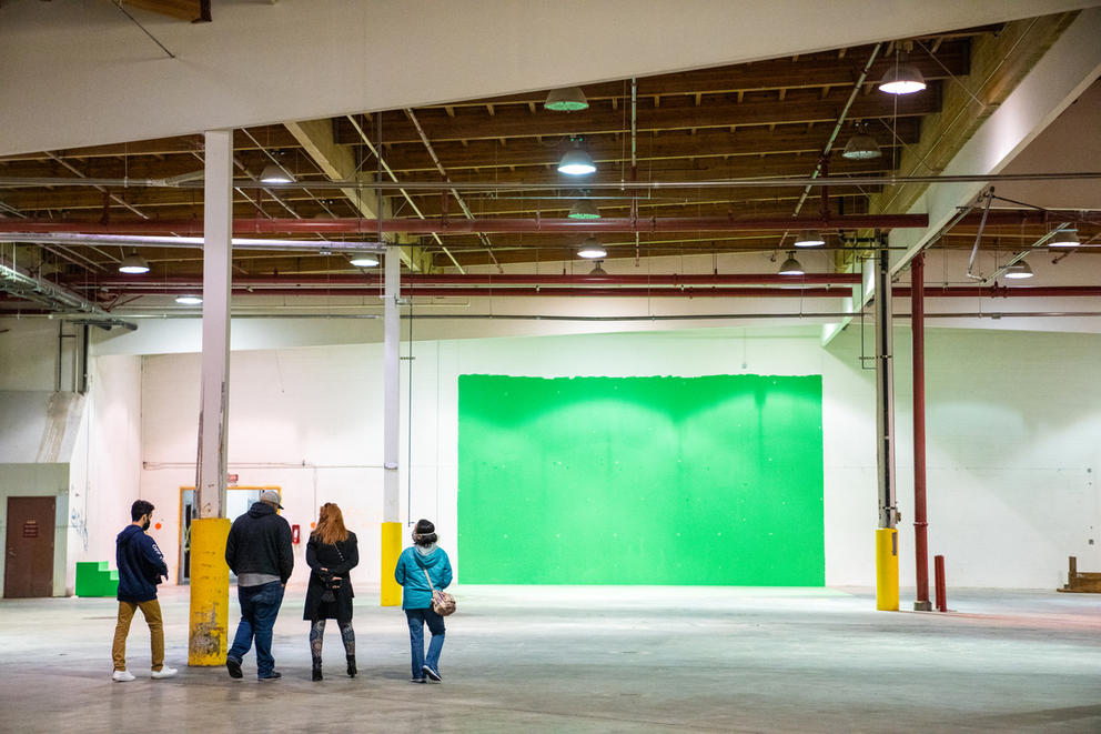 Four people stand in a large room with beams, in the back a green square is painted on a white wall
