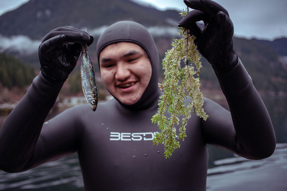 A snorkeler in a full-body wetsuit holds up a fish and a plant