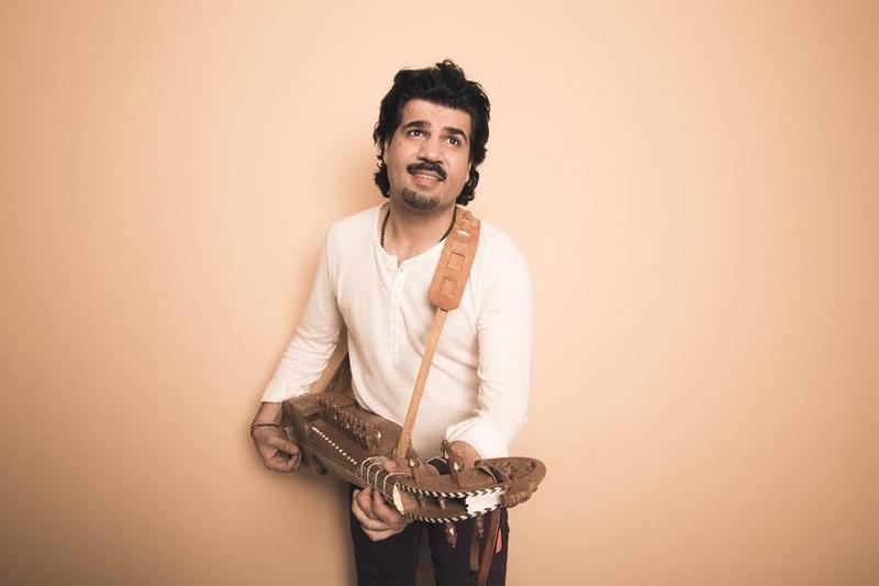 A man with black hair and a mustache stands in front of a peach backdrop and is wearing a long sleeve white shirt. He is holding a rubab instrument.