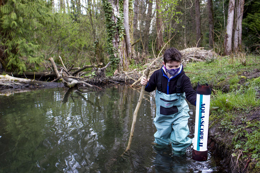 a young boy wearing waders and holding a walking stick and aquascope wades through a pond