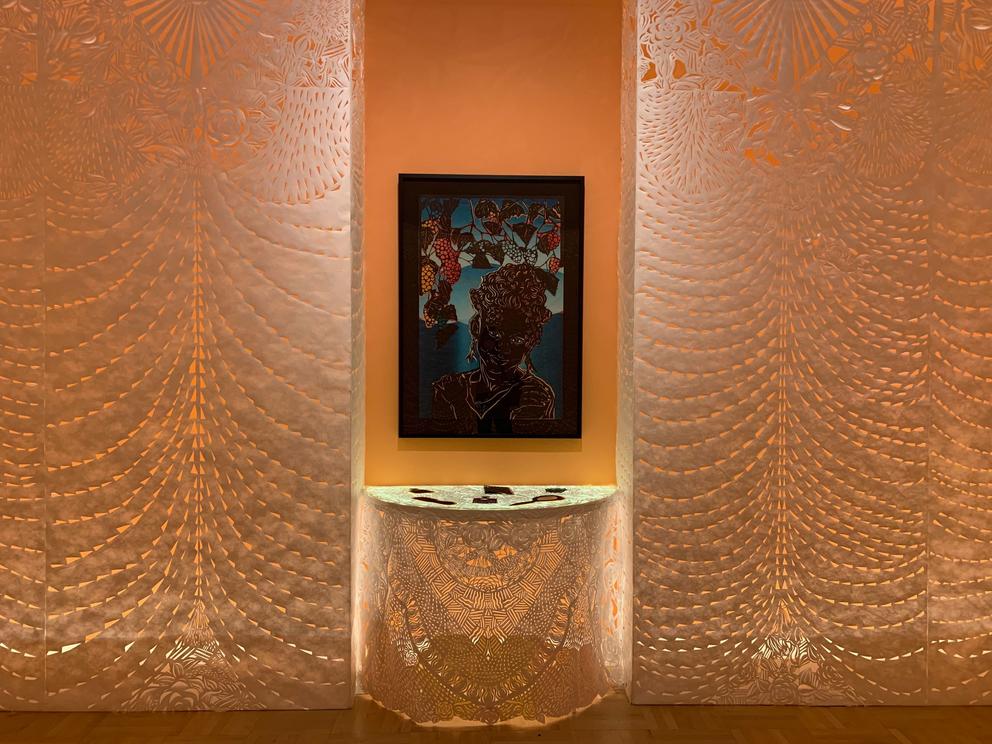 An altar from cut paper surrounded by two columns of cut paper. A painting hangs in the alcove in the middle.