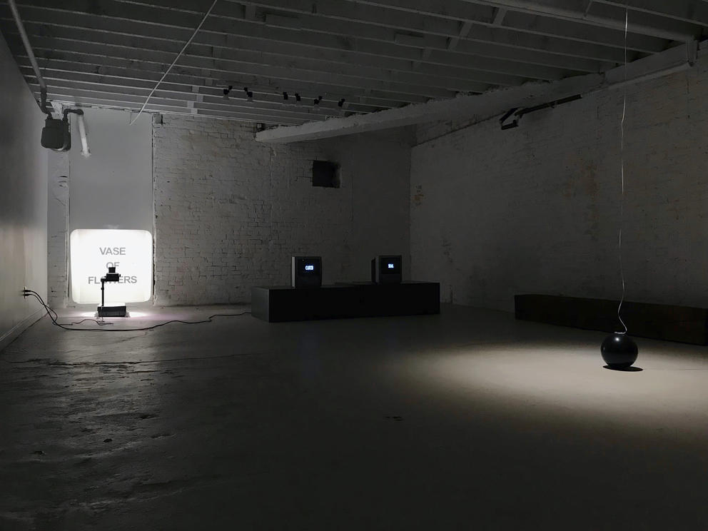 A dark, sparsely lit space featuring a projector in the left hand corner and a black bowling-like ball in the right corner