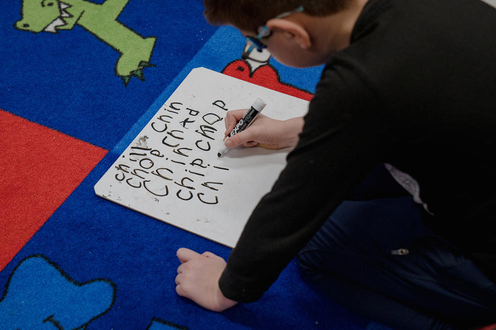 A student sits on the floor writing on a white board. The words are: chill, chin, chop, Chad and chip.