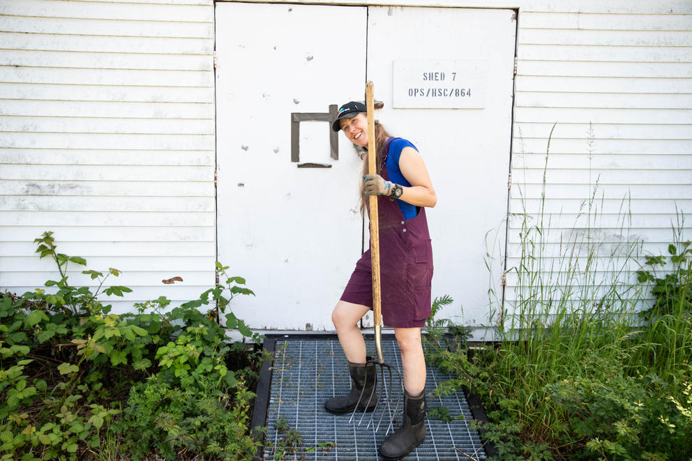 A person in purple overalls with long hair closes the door to a white shed while holding a shovel. 