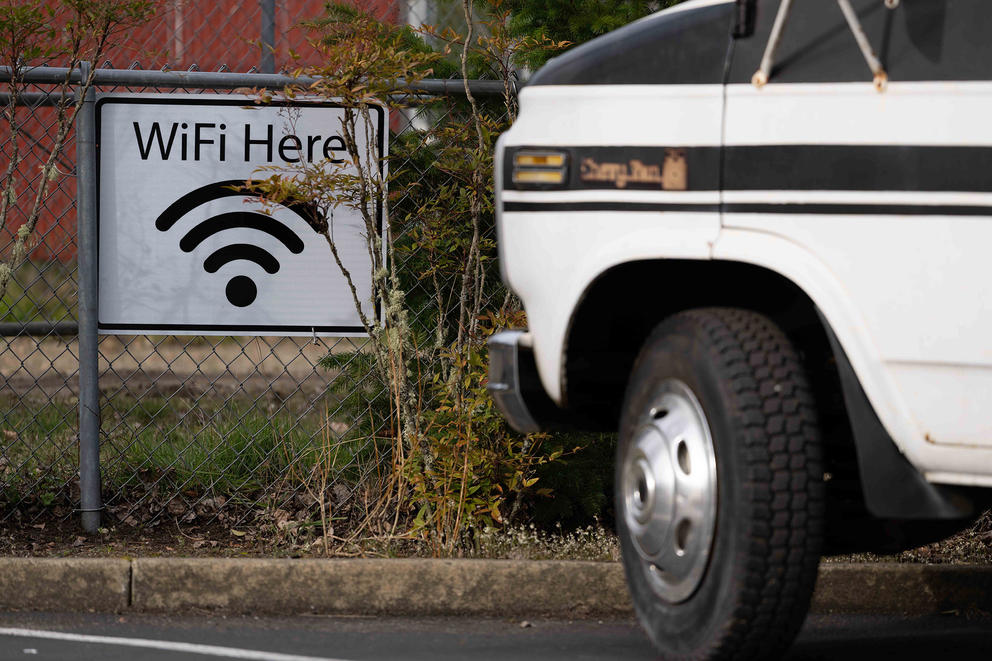 A sign reading Wi-fi here hangs on a chainlink fence in parking lot.