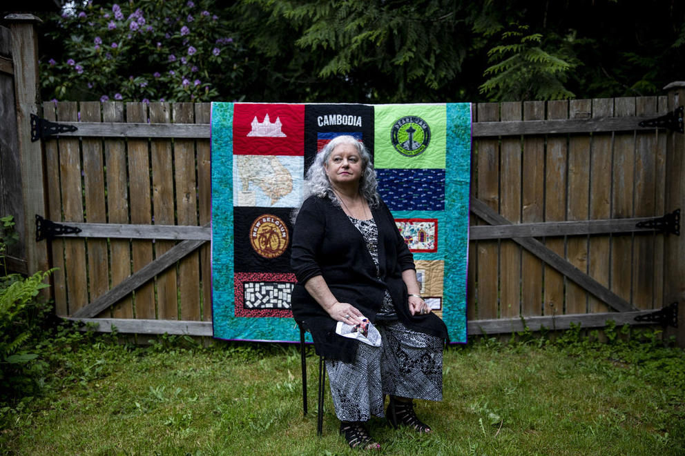 A seated woman poses in her yard in front of a quilt hung on the fence behind her