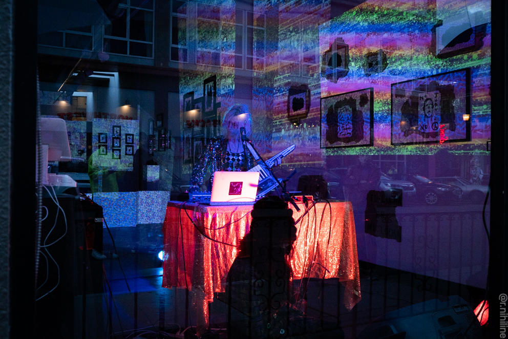Person in glitter vest behind laptop, which is sitting on a table covered with a sparkly orange drop cloth. Reflections of buildings are seen in the windows, and behind the singer projections illuminate white walls covered in paintings.