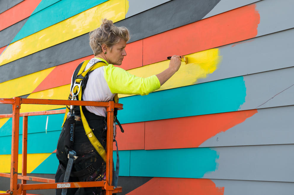 a woman in overalls painting a yellow orange and turquoise stripes on a building