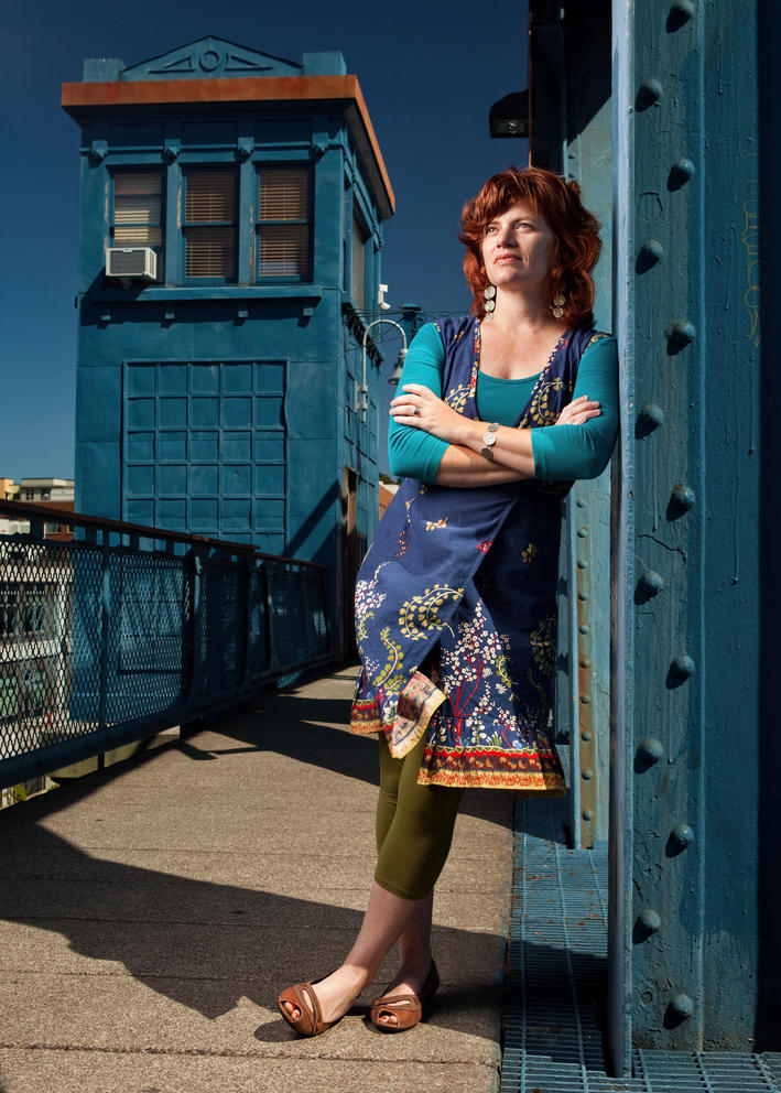a woman with red hair wearing a turquoise top and blue dress stands with arms crossed near a blue bridge tower