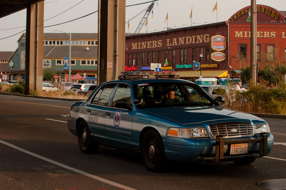 A police car drives along the Seattle waterfront with the Miner's Landing building in the background