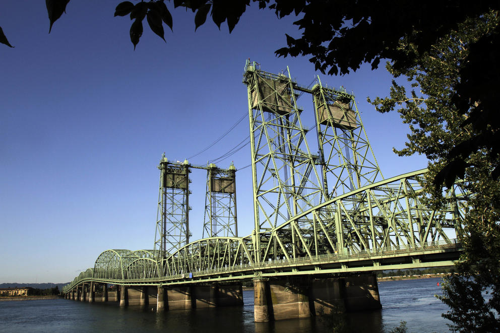A photo of the green Interstate 5 bridge over the Columbia River, with trees framing the right of the frame and blue sky behind