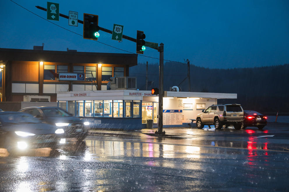 A small burger restaurant is lit up at night. It is raining and the rain drops are illuminated by the headlights of passing cars.