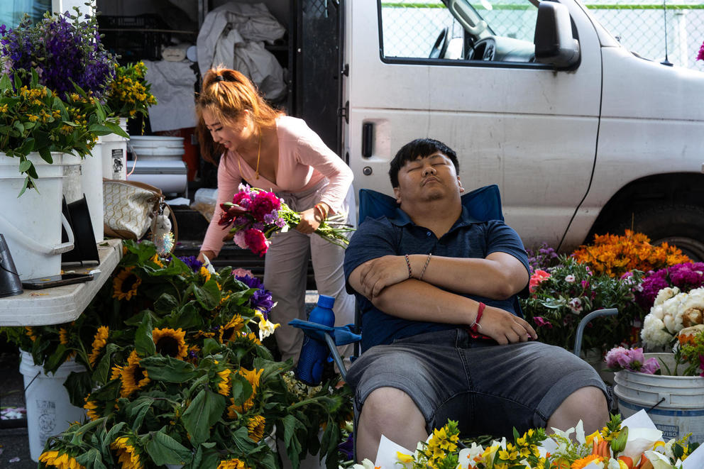 man sits in a chair napping while a woman selects flowers to add to a bouquet from buckets sitting outside a van