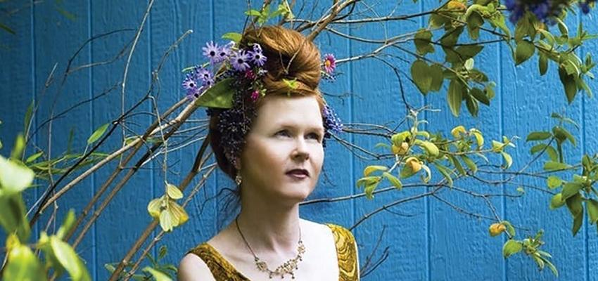 Sarah Cahill stands in front of a blue background within branches of leaves, her red hair interwoven with flowers in a bun on top of her head.