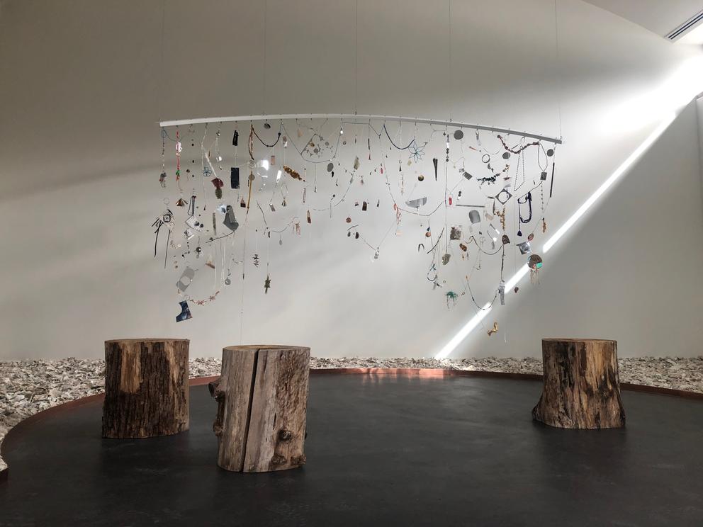 tree stumps on darker floor, surrounded by a oyster shell floor and a mobile that consists of a weaving of various materials, including pieces of copper, styrofoam, beadwork and more.