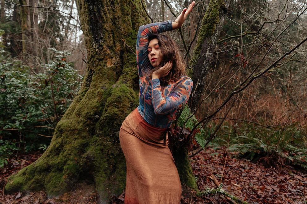 Person in orange skirt in front of tree with moss, in dancing position