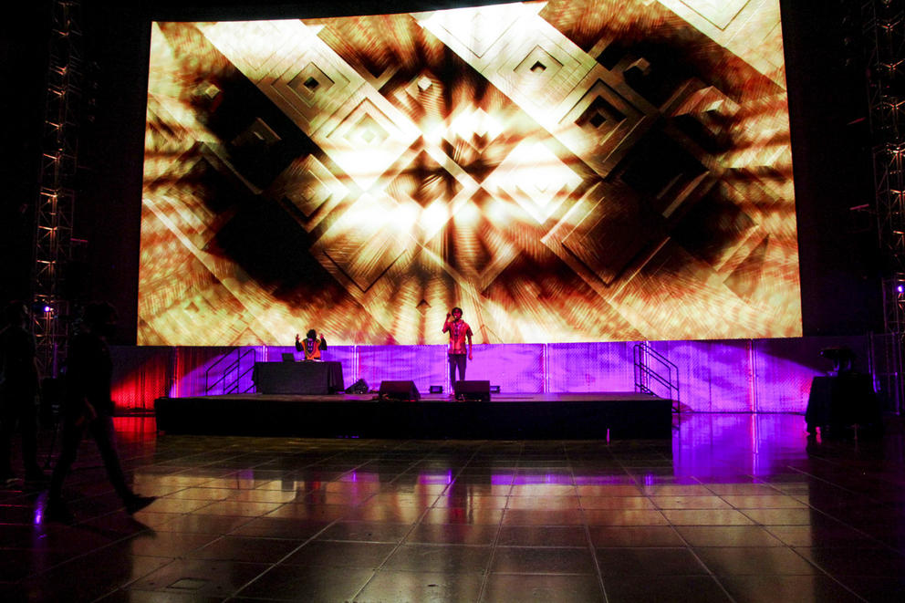 stage in empty room, tiles reflect the light of a large LED screen in the back that shows purple and golden art. One performer is on stage.