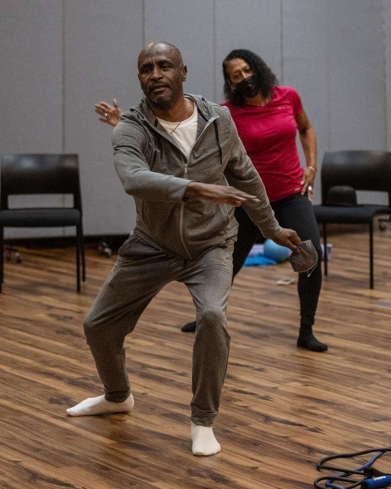 A man wearing gray and a woman in hot pink dance on a hardwood floors of a dance studio