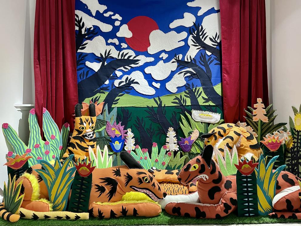 Animal puppets sit within exotic plants in front of a blue sky background with clouds — all made out of felt.