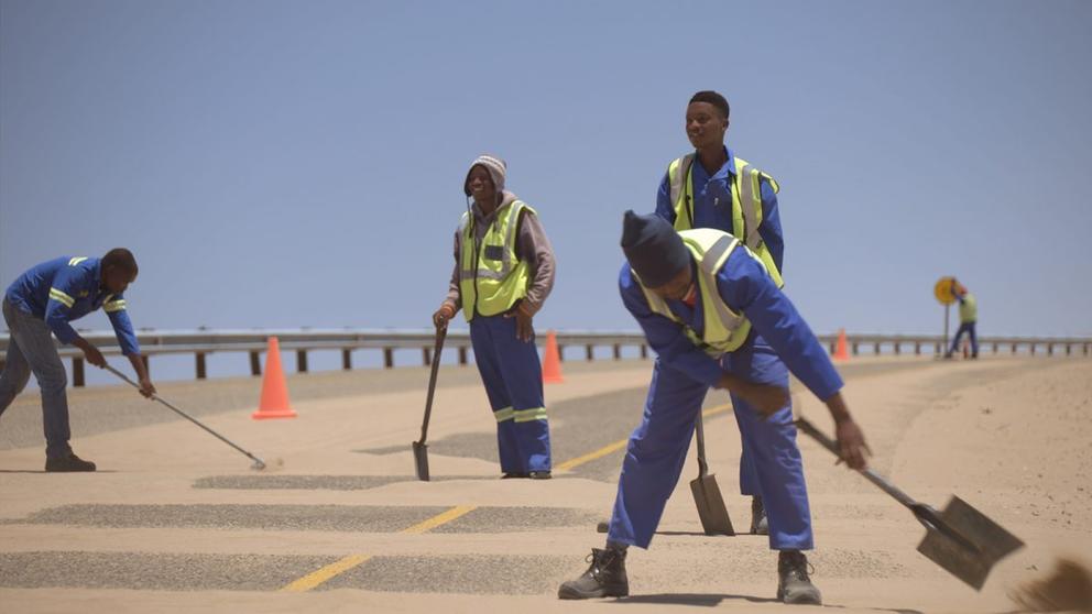 men wearing safety vests sweep sand off a road