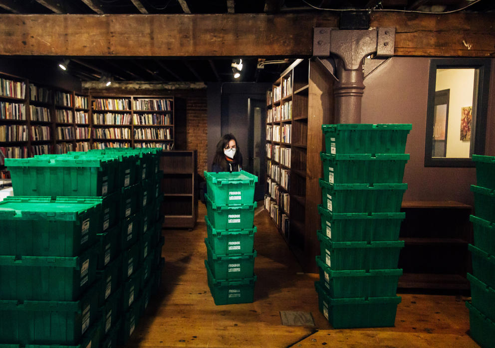 Person in dark store, painted purple and featuring large beams and cast iron columns, drags a stack of green boxes. The person is masked.