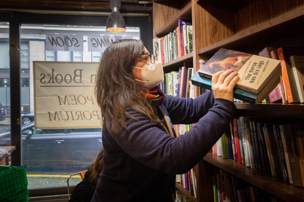 Person in dark blue sweater, dark long hair and white mask places a few books in wooden shelving unit.